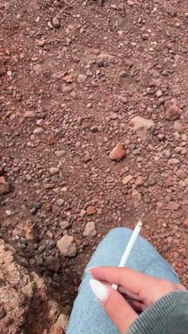 A hand with long, pointed fingernails holds a lit cigarette against a background of red gravel, then moves the cigarette out of view. The gravel area leads to a dark, barren landscape, beyond which a distant, scenic view of a town or city is visible beneath a partly cloudy sky.