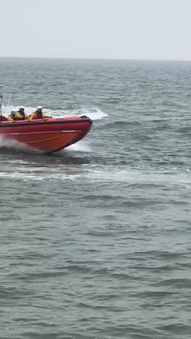 A red inflatable boat with four people wearing yellow jumpsuits and white helmets is moving at high speed through the water. The boat has "RNLI" printed on its side. A person with gray hair is seen from behind, taking a picture of the boat with a smartphone. The boat passes a pier with people walking on it. The sky is overcast.
