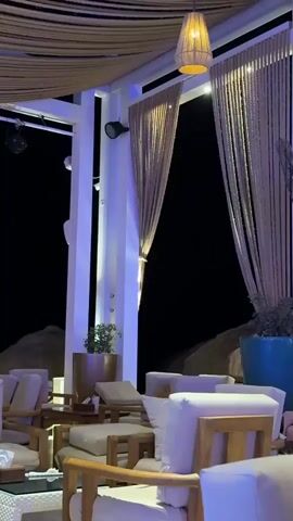 The video shows an outdoor seating area at night, possibly a restaurant or lounge. Several white upholstered chairs and wooden tables are arranged under a canopy. Draped curtains made of light-colored rope or fabric hang from the canopy. Blue and white lights illuminate the space. A large, industrial-style fan is mounted on a white pillar. In the background, a dark sky is visible, with what appears to be a sandy or rocky incline. A person with red hair is seated at one of the tables, holding a phone to their ear. The camera pans across the scene, focusing on the seating, the fan, and briefly on the person.