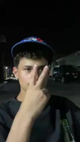 The video shows a young man standing in front of a building at night. The man is wearing a black shirt. He is smiling. In the background, there is a dark-colored car parked. Another young man with curly hair is also visible. The surroundings appear to be in an urban area.