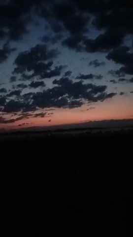 This is a video of a dusky landscape. The sky is a deep blue with scattered clouds that appear dark. A thin band of reddish-orange light is visible at the horizon, indicating a sunset or sunrise. The foreground is a dark, silhouetted area, possibly a field or open ground, with a faint haze or mist visible just above it. There are no people or identifiable objects in the video. The scene appears to be outdoors and is dimly lit.