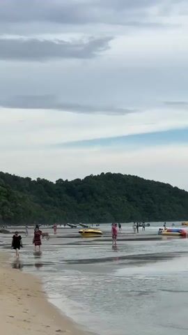 A video pans across a wide, wet sand beach with calm, shallow water lapping at the shore. In the foreground, small waves are rolling in. In the mid-ground, several people are visible, some walking on the wet sand, others wading in the shallow water. There are also several colorful inflatable tubes and a yellow banana boat on the sand and in the water, suggesting recreational activities. In the background, a lush green, tree-covered hill forms a backdrop against a cloudy sky with patches of blue. The overall scene depicts a moderately populated beach on a partly cloudy day, with a focus on the sandy shore, shallow water, and the natural landscape.