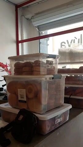 A stationary shot showcases stacks of clear plastic bins filled with pastries. The bins are arranged in a multi-tiered display on a concrete-like surface. The pastries appear to be a variety of rolls, donuts, and croissants. The bins closest to the camera show labels and branding stickers. A black shoulder bag rests on the concrete surface next to the bottom bin. In the background, a partially-open roll-up metal door reveals a glimpse of the outside, where a sign with the text "salvi..." is visible, though the complete word is not clear. The color scheme is predominantly neutral, with the pastries and bins being in shades of brown and white. The environment appears to be a bakery or food preparation area.