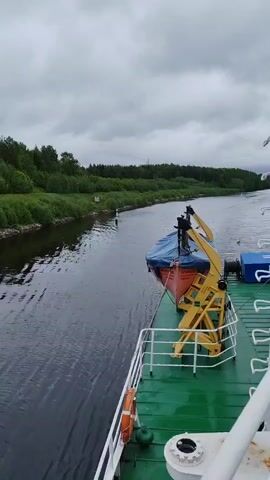 The video shows a view from the deck of a boat or ship traveling down a waterway. The water is dark and reflects the cloudy sky. Lush green vegetation lines both sides of the waterway. A small, white navigation marker stands on the left bank. On the ship's deck, an orange lifeboat, covered with a blue tarp, is held in place by a yellow crane. White railings surround the deck, and a green surface covers the deck.