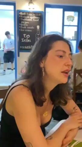 Here is a concise description of the video:

The video features a woman with dark hair, wearing a black tank top and earrings, speaking while seated at a table inside what appears to be a restaurant. The restaurant's decor includes blue and white accents, and a chalkboard menu displaying Greek food options is visible in the background. Other people are present in the restaurant.