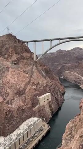 The video shows a panoramic view of the Hoover Dam and the Colorado River. The camera pans from left to right, revealing the immense scale of the dam and the surrounding landscape. In the foreground, the top of the dam is visible, with its intricate electrical infrastructure. The dam itself is constructed of concrete and spans a wide canyon, holding back a vast body of water, Lake Mead. The water appears dark and calm, reflecting the overcast sky.

Further back, nestled into the cliff face, is a white, rectangular building with arched windows. This is likely one of the powerhouses of the dam. The surrounding mountains are rugged and arid, characterized by reddish-brown rock formations and sparse vegetation.

Dominating the upper part of the frame is a large, modern bridge, the Mike O'Callaghan–Pat Tillman Memorial Bridge, which carries traffic over the Colorado River. Its elegant arch design stands in stark contrast to the natural ruggedness of the canyon. The bridge's concrete piers rise majestically above the dam and the canyon walls. The sky is overcast, creating a diffused light that casts a soft glow over the scene. Power lines can be seen strung between tall metal towers on the mountain ridges, indicating the dam's role in power generation and distribution. The video captures the engineering marvel of the Hoover Dam in its dramatic natural setting.