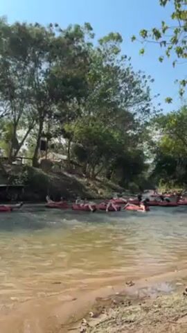 A group of people are leisurely floating down a shallow river in red inflatable tubes on a sunny day. The river is surrounded by lush green trees, and there is a wooden structure with steps on the riverbank. The water is clear and the river bottom is visible. One of the tubes holds four people, two women and two men, who are holding a small bucket. One of the men is wearing sunglasses and appears to be enjoying the float. There is a tire on the bank.