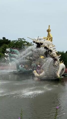 A multi-headed dragon fountain is active in a pond. The dragon is a sculpture made of white and green sections with golden accents. Water sprays from various points on the sculpture, creating a mist and a cascading effect into the pond. In the background, there are green trees and a golden-roofed pavilion. The sky is overcast. There are no people visible in the immediate vicinity of the fountain.