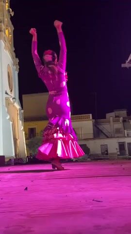 A woman in a purple polka dot dress with ruffled pink trim dances on a pink stage at night. Her arms are raised above her head, and her body is arched backward. The stage is set against a backdrop of a building with arched windows and a dark night sky.