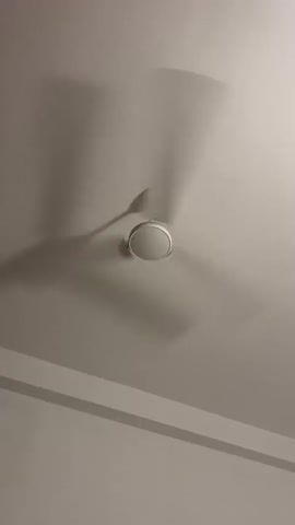 This media file shows a white ceiling fan rotating against a white ceiling. The fan blades are blurred due to their motion, casting faint shadows on the ceiling. Below the fan, a horizontal band, likely a molding or a beam, runs across the scene, creating a dividing line between two slightly different shades of white. The lighting suggests an indoor setting, possibly daytime given the natural light implied by the shadows. There are no people or explicit text visible. The overall impression is one of a typical domestic or office environment in Kadewatta, Sri Lanka, where a ceiling fan is in operation, likely to provide cooling or air circulation. The scene is static, focusing solely on the fan's movement and the surrounding architecture.