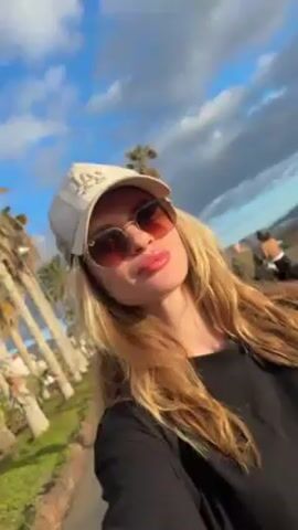 A woman is taking a selfie outside with a slightly angled, upward shot. She is wearing a black shirt, a light-colored baseball cap with a logo, and sunglasses. Her long, blonde hair is wavy. The background includes palm trees, green grassy areas, and a cloudy blue sky. There are other people in the background, but they are out of focus. The camera appears to be hand-held.