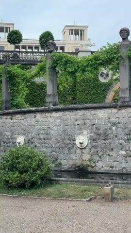 A panning shot reveals a stone wall with decorative lion-head fountains, flanked by manicured hedges. Above the wall, two stone pillars topped with busts of men are entwined with green vines. Further back, an arched entryway is visible, also covered in greenery. In the background, a stately building with a colonnaded facade stands against a cloudy sky, and mature trees provide a lush canopy.