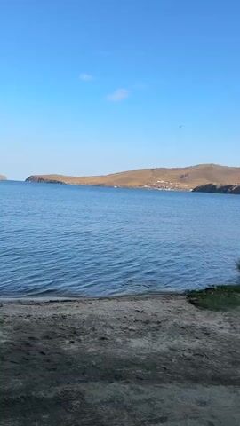 A tranquil blue lake is bordered by a sandy shore in the foreground. Beyond the water, rolling hills with sparse vegetation and a small village dot the landscape under a clear blue sky. A few boats are visible on the water in the distance.