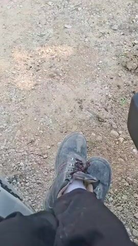 The media file shows a person's legs and feet, wearing dark pants and worn, grey work boots with red laces. The person is seated, with their legs crossed at the ankles. The ground beneath them is covered in gravel and dirt, suggesting an outdoor, possibly rural or construction setting. The lighting indicates it is daytime, with sunlight casting shadows. There is no visible text in the media file. The overall scene is simple and focuses on the footwear and the immediate surroundings. The boots appear to have seen significant use, suggesting the person might be engaged in physical labor or outdoor activities. The location context provided is Hîncești, Moldova.
