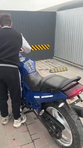 A person stands next to a blue motorcycle, repeatedly attempting to start the engine. The motorcycle has "Suzuki Motorsport" written on the body. It is parked on a brick-paved area next to a gray corrugated metal wall with a black and yellow striped barrier. The motorcycle appears to be damp.