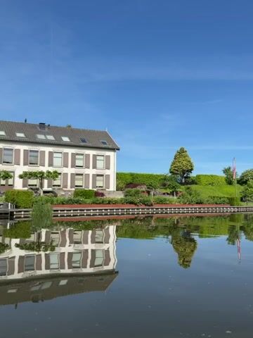 The camera is on a boat moving slowly on a canal on a bright, sunny day. The water is calm and reflects the buildings and trees on the canal banks. The sky is a clear blue with a few wispy clouds. Several houses are seen on the banks of the canal, each with well-maintained gardens and lawns. A flag is flying in the distance. The boat is moving slowly, and the camera remains still, capturing the peaceful scenery as it passes by.
