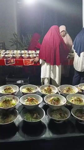 The video shows an outdoor scene at night, possibly a buffet or event. In the foreground, there are multiple bowls of food, likely soup or noodles, arranged on a dark surface. The bowls are white and contain a light-colored broth with visible ingredients like noodles, meat, and vegetables, topped with fried onions and green herbs. In the background, three women are standing behind a long table covered with a red tablecloth. The table is laden with more bowls of food and some boxes or containers. The women are dressed in hijabs and traditional clothing. One woman in a maroon hijab is reaching towards the table, appearing to be serving or arranging food. Another woman in a light beige hijab is looking down at the table, and the third woman on the right, wearing a dark blue hijab, is also facing the table. The environment appears to be outdoors with some greenery visible behind the table, illuminated by artificial lighting, creating a slightly blurred background. The overall impression is a communal meal being prepared or served.
