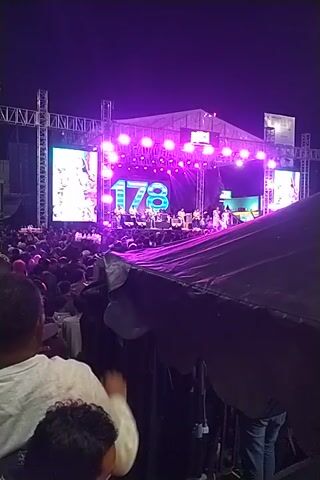 Here is a description of the video:

The video captures a live performance on a large outdoor stage at night. The stage is brightly lit with pink and white lights, and two large screens flank the central performance area. On the screens, images of the performing band are displayed, and the number "87" is prominently featured above the musicians. A large crowd is visible in front of the stage. 

The perspective of the video is from within the audience, where a person is holding up a black object, possibly a tarp or some sort of makeshift cover, partially obstructing the view of the stage. The person's arm and hand are visible, along with the heads of other audience members. The video is shot vertically and appears to be taken with a mobile device.