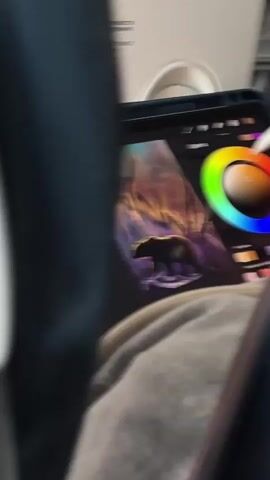 The video shows a close-up view of a person's hands using a stylus on a tablet computer. The tablet displays a digital painting of a bear walking through a forest. A color wheel is visible on the right side of the screen, and the stylus is being used to select a color. The person is wearing a gray hooded sweatshirt, and the background appears to be the interior of an airplane or train.