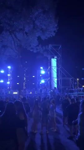 A large outdoor crowd is gathered at night in front of a stage. The stage has several scaffolding structures with large speakers. Blue lights illuminate the stage and parts of the crowd. People are standing and talking, some are looking towards the stage. The atmosphere appears to be that of a concert or festival.