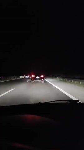 A black car is driving on a highway at night, with its taillights illuminated. There are white lines on the road indicating lanes. Another car is visible in the distance ahead of the black car. The sky is dark and there are no other visible surroundings. The camera appears to be inside another vehicle following the black car.