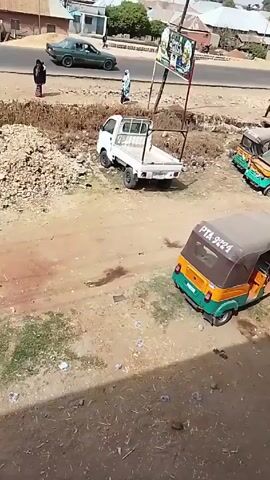 The video shows an urban landscape. In the foreground, there is a dirt area with three auto rickshaws parked, two of which are visible in detail; they are orange and green, and one has a license plate reading "PTA-9224". A small white pickup truck is parked nearby, facing away from the camera. Behind the truck, there's a metal frame holding up a vertical sign with advertisements. A road runs parallel to the parked vehicles, with a green sedan driving on it. Two people are walking along the side of the road. Buildings and trees are visible in the background. The scene is sunlit. Litter is scattered across the ground. The video seems to be taken from an elevated position, potentially from a building.