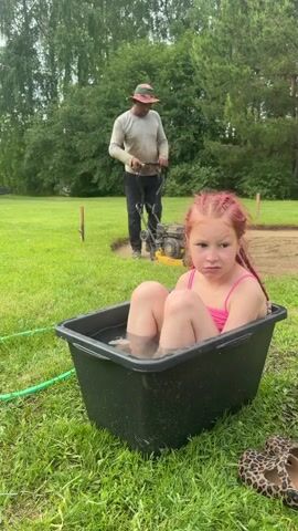 A young girl with red braided hair and a pink swimsuit sits in a black plastic tub filled with water on green grass, looking towards the right side of the frame. In the background, a man wearing a hat and light-colored shirt operates a yellow plate compactor within a circular excavated area. Trees are visible in the far background. A pair of leopard-print shoes rests on the grass next to the tub, and a green hose is also visible on the ground. The girl first looks towards the man, then turns her gaze forward.