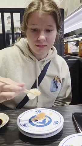 A young woman with blonde hair is eating sushi at a restaurant. She is wearing a light-colored hooded sweatshirt with a cartoon graphic of two people on the front. She is holding chopsticks in her right hand and is bringing a piece of sushi to her mouth. In front of her is a white plate with a blue logo and a piece of sushi. There is also a small white dish with soy sauce to the left of the plate. The background is blurred but appears to be the interior of a restaurant with dark wooden tables and some shelving.