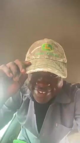 The video is a selfie-style shot of a man wearing a camouflage baseball cap with the John Deere logo, a light blue button-down work shirt, and a black t-shirt underneath. He is smiling and looking directly at the camera. The man sticks his tongue out and places a small, hard-boiled egg on it. The background is blurry but appears to be the interior of a vehicle with a tan or light-colored ceiling. There is bright light coming from an unseen source, possibly the sun or an interior light.