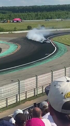 In this video, three cars are drifting around a corner on a race track, producing a large amount of smoke. The cars are close together, with the car on the left being dark colored with pink and white accents, the car in the middle being white with green and black accents, and the car on the right being white. The race track is paved and has green painted curbs. Beyond the track, there is a grassy area with trees in the background. In the foreground, a crowd of people are watching the event from behind a fence. The sky is blue with some clouds. The camera angle is from above and slightly behind the spectators.