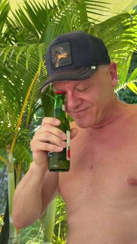 A man with a bare chest stands in front of palm fronds, holding a green beer bottle up to his eye. He is wearing a baseball cap with a dog on it. He appears to be looking through the bottle.