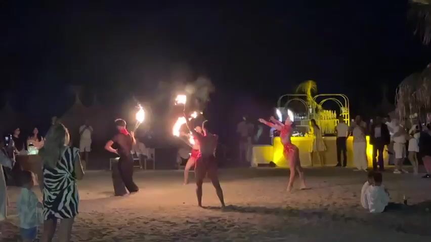 A group of performers are engaged in a fire dancing show on a sandy beach at night. Three performers are actively wielding torches that are aflame. One performer, dressed in dark pants and a red shirt, is on the left, twirling a torch. Another performer, in red attire, is in the center, holding torches at chest level. A third performer, also in red, is on the right, with a torch held low. The background shows an audience of people, some sitting and some standing, observing the performance. There is also a structure resembling a bar or stage with decorative elements and lighting. Palm trees are visible on the right side of the frame. The overall setting is a beach at night, illuminated by artificial lights and the fire from the torches.