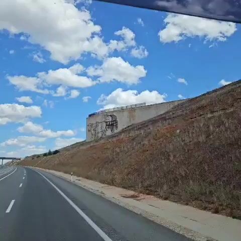 The video shows a view from inside a vehicle, looking out at a mural on a concrete wall. The mural depicts a horse-drawn wagon being pulled by two oxen, with a person driving the wagon. The wall is situated on a grassy embankment next to a highway. The sky is blue with scattered white clouds. The camera is moving, suggesting the vehicle is in motion.