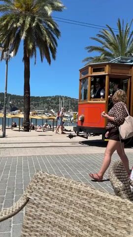A vintage wooden tram car passes a café on a sunny day in a tropical locale. Palm trees line the street, and people are visible on a beach in the background. The tram is orange and brown, with passengers visible through the windows. A woman in a polka dot shirt and shorts walks alongside the tram as it moves. The foreground shows part of a wicker chair, suggesting the perspective is from someone seated at the café. The sky is clear blue.