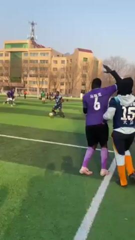 The video shows a soccer game in progress. People in green, white, and purple jerseys are running on a green field, kicking a soccer ball. Two men in purple and blue tracksuits are standing on the sidelines, watching the game. A tall building is visible in the background. The sky is blue and the sun is shining.