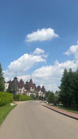 This is a vertical, medium-shot video recording of a picturesque setting on a bright, sunny day. The scene depicts a grand, ornate building with a distinctive architectural style, reminiscent of a castle or a large manor. The building features multiple spires, dark brown roofs, and light-colored walls, with what appear to be flags or weather vanes atop some of the spires. It is flanked by lush green trees and a meticulously maintained hedge that lines a paved road.

The road curves gently, leading the viewer's eye towards the building. On the right side of the road, there's a well-kept pathway and some decorative elements, including large, rounded, dark-colored objects that could be large stones or sculptures, nestled amongst greenery. The sky is a vibrant blue, dotted with fluffy white clouds, indicating fair weather. The overall impression is one of serene beauty and a touch of fairytale charm.

The video appears to be taken from a moving vehicle, likely a car, as evidenced by the slight blur and the perspective of the road. The time of day is clearly daytime, given the bright sunlight and the clear visibility. No people are visible in the frame. The location is identified as Molokovo, Russia. The mood is peaceful and visually appealing, showcasing the grandeur of the architecture and the natural beauty of the surroundings. There is no visible text in the media file.