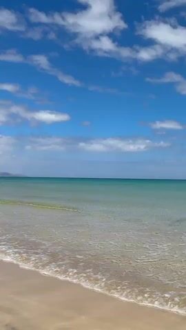 The video shows a beach scene on a sunny day. The sky is blue with scattered white clouds. The ocean water is clear and turquoise, with gentle waves lapping onto the sandy shore. People are seen swimming and enjoying the water. A jet ski is visible in the distance. The camera pans along the beach, revealing a rocky outcrop with a building on top and then a stretch of sandy beach with people sunbathing and surfing. In the background, there's a building on a hill and a trailer carrying surfboards. The overall impression is that of a relaxing and pleasant day at the beach.