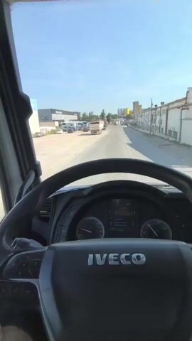 The video shows a first-person perspective from inside the cab of an IVECO truck driving on an asphalt road. The driver is holding the steering wheel.  The truck is moving slowly on a sunny day. Other trucks and parked vehicles can be seen along the side of the road, as well as warehouse-like buildings. The side mirrors of the truck are visible and show other vehicles on the road. The dashboard of the truck is visible, showing the IVECO logo.