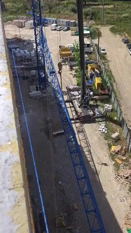 A construction site with two cranes. One crane, a blue lattice boom crane, is partially visible on the left. The second crane, a mobile crane with a black boom, is on the right. The mobile crane's boom is extended and a hook is visible near the end of the boom. There are stacks of construction materials, including concrete blocks and yellow metal beams, on the site. The ground is covered in gravel and dirt. There are a few vehicles parked in the background. The scene appears to be under construction during daylight.