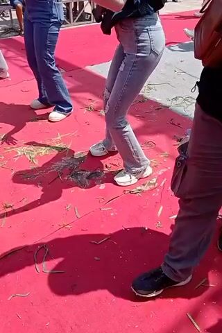 A group of individuals are dancing on a red carpet outdoors. The carpet is somewhat worn and has scattered green debris on it. The people are wearing casual clothing, including jeans and t-shirts. One person in the foreground is wearing a black t-shirt and grey cargo pants, facing away from the camera. Behind them, two women are dancing. The woman on the left wears dark blue jeans and a black and blue shirt. The woman on the right wears ripped light blue jeans and a similar black and blue shirt. Both women have their hair down. In the background, other people and structures are visible, suggesting an event or performance is taking place. The lighting suggests it is daytime.