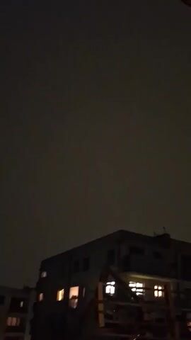 A nighttime video captures a low-angle view of a dark, overcast sky above several buildings. The buildings are dimly lit from within, with warm yellow and orange light emanating from some windows. The perspective suggests the camera is positioned on a lower level, looking upwards. The scene is dark and still, with no visible movement or sound. The buildings appear to be apartment complexes or similar multi-story structures.
