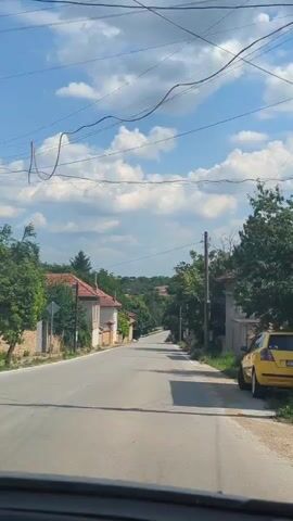 The video shows a road in a rural village on a sunny day. The road is paved and appears to be empty of people. On either side of the road are houses with red-tiled roofs, fences, and trees. The sky is blue with white clouds. The camera is mounted inside a vehicle, suggesting the video is taken from a moving car.