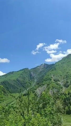 The video shows a bright, sunny day with a clear blue sky dotted with white clouds. Lush green trees and vegetation dominate the foreground. In the background, a steep, verdant mountain slope rises, with patches of snow visible near the peak. The camera appears to be moving, possibly indicating that the video was shot from a vehicle or while walking.