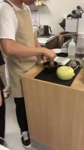 A person in a tan apron is preparing matcha. The person is using a chasen whisk in a black matcha bowl to mix a green liquid. The bowl is placed on a black mat on a light-colored countertop. Next to the bowl is a yellow teapot. Behind the person is a white refrigerator, a coffee machine, and other kitchen equipment.