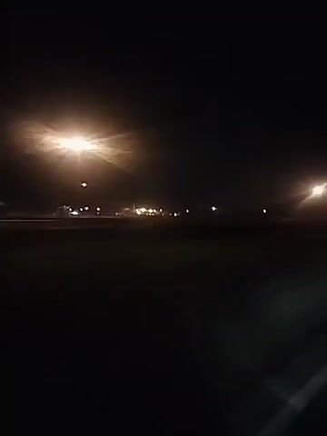The video shows a view from a vehicle moving along a road at night. The road is paved with a white line marking the edge. In the distance, a series of street lights illuminate a flat, open area. The video is slightly shaky, giving the impression of movement. The overall lighting is dark, with the street lights being the primary source of illumination.