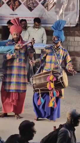 A man in a blue turban and a colorful plaid kurta plays a dhol drum. Beside him, another man in a red turban and matching plaid kurta plays a flute. A third man, to the left, wears a blue kurta and a green shawl. They appear to be performing for an audience. Behind them, a banner displays images of traditional Indian art and calligraphy. The ground is a mix of dirt and pavement.