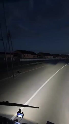 The video captures a point-of-view shot from inside a vehicle at night, likely a truck, as indicated by the wide windshield and dashboard view. Bright headlights illuminate the empty asphalt road ahead, which has a white line marking its center. In the background, a row of houses with illuminated windows lines the left side of the road. The sky is dark blue with some clouds, suggesting dusk or early night. The vehicle is moving forward, with the scenery subtly blurring due to motion. A small electronic device, possibly a dashcam, is visible on the dashboard. The overall atmosphere is one of solitary travel through a quiet, dimly lit suburban or rural landscape.