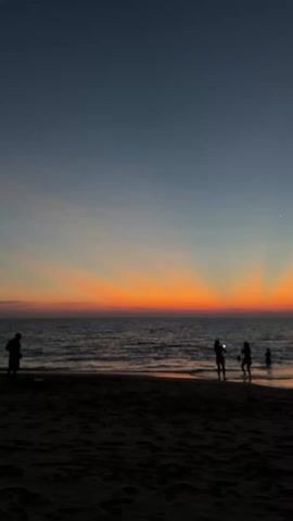 The video depicts a picturesque sunset at the beach, with a dark blue sky transitioning to a vibrant orange horizon where the sun has set. Silhouetted against this backdrop are three figures: two individuals standing close together at the water's edge, possibly a couple, and a child slightly further out in the water. The foreground is composed of dark sand, contrasting with the illuminated ocean and sky. Gentle waves are visible, and a single bright star or planet is discernible in the upper right portion of the sky. The scene conveys a sense of tranquility and natural beauty.