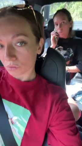 In a daytime video, two women are inside a car. The woman in the foreground, wearing a red t-shirt with a graphic design and sunglasses on her head, records a selfie video. She points a finger backward towards the second woman. This second woman, wearing a black long-sleeved shirt, is seated in the back and is actively talking on a mobile phone. The car appears to be in motion, indicated by the blurred trees and greenery visible through the windows, suggesting an outdoor, possibly rural or suburban setting. The woman in the foreground has green eyes and light makeup, showing a slight smile. The overall mood is casual.