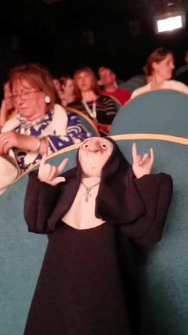 A low-angle, slightly shaky shot captures a peculiar scene in a theatre. In the foreground, a doll dressed as a nun, complete with a black habit, white collar, and a silver cross necklace, sits prominently on a teal-colored seat with gold piping. The doll is making the "rock on" hand gesture with both hands, its face showing a rather grotesque appearance with exaggerated features. Behind the doll, the shot is filled with a number of blurred figures, presumably other audience members. These appear to be mostly women, some of whom are wearing glasses or have light-colored tops. The background is dark, as expected in a theater setting during a performance, with a single, faint light source visible above and behind the audience. The overall atmosphere is one of incongruity, the sacred figure of the nun juxtaposed against the rebellious "rock on" gesture, creating an absurd and humorous effect.